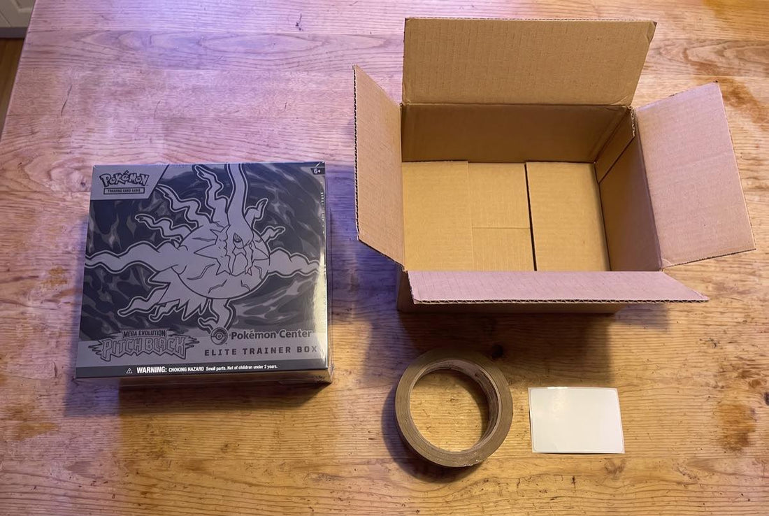 Sealed Pokemon Pitch Black Elite Trainer Box on a table beside an open cardboard shipping carton, parcel tape and a blank postage label
