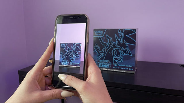 A sealed Phantasmal Flames Pokemon Center Elite Trainer Box in a clear case, photographed with a phone on a desk