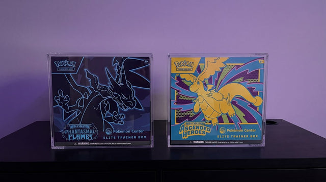 The Phantasmal Flames and Ascended Heroes Pokemon Center Elite Trainer Boxes in clear display cases on a black desk
