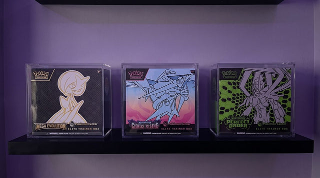 Three sealed Pokemon Center Elite Trainer Boxes standing upright in clear acrylic cases on a black wall shelf